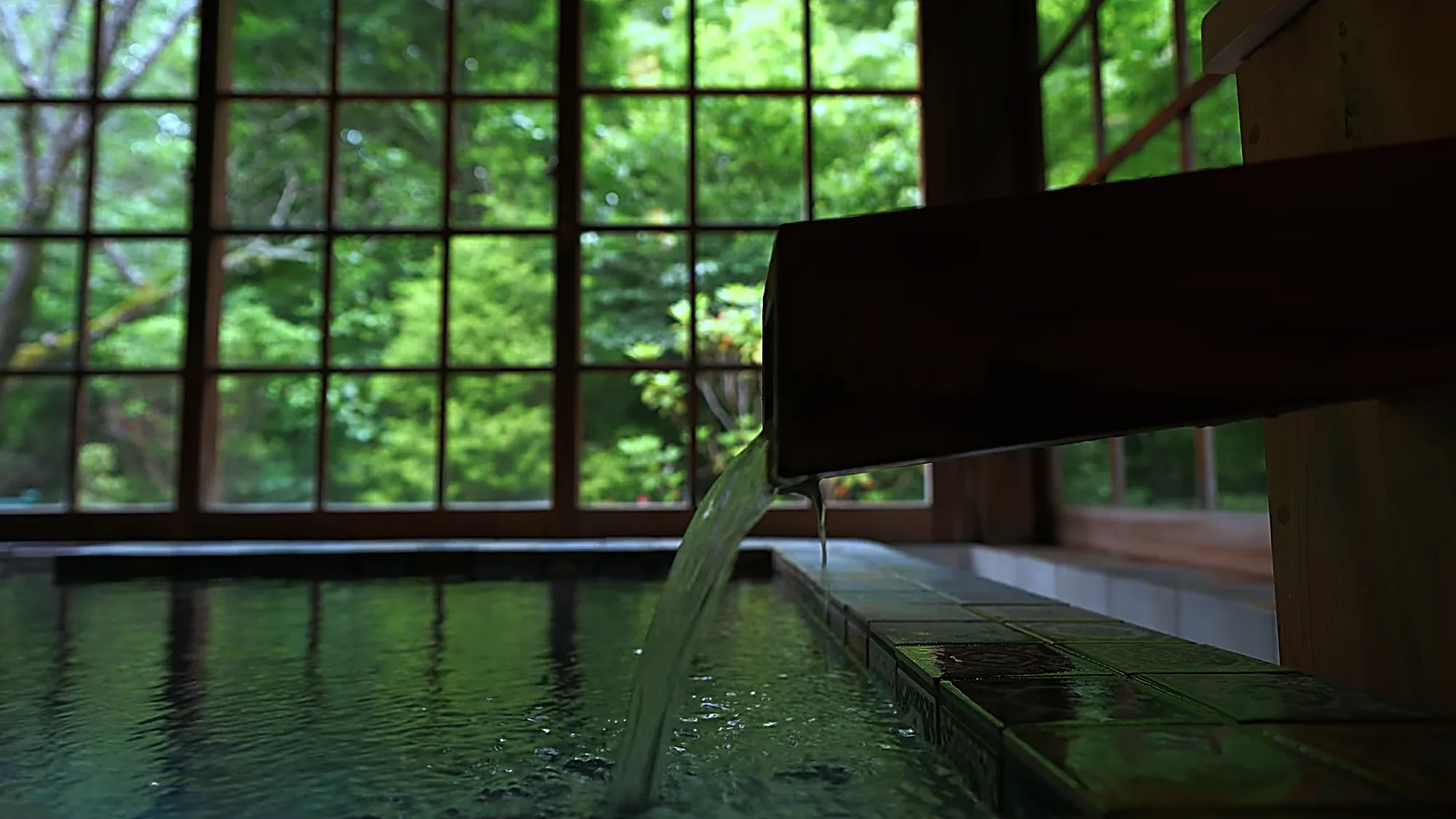 An indoor bath at Kurokawa Onsen fed by a timber spout, windows looking onto green