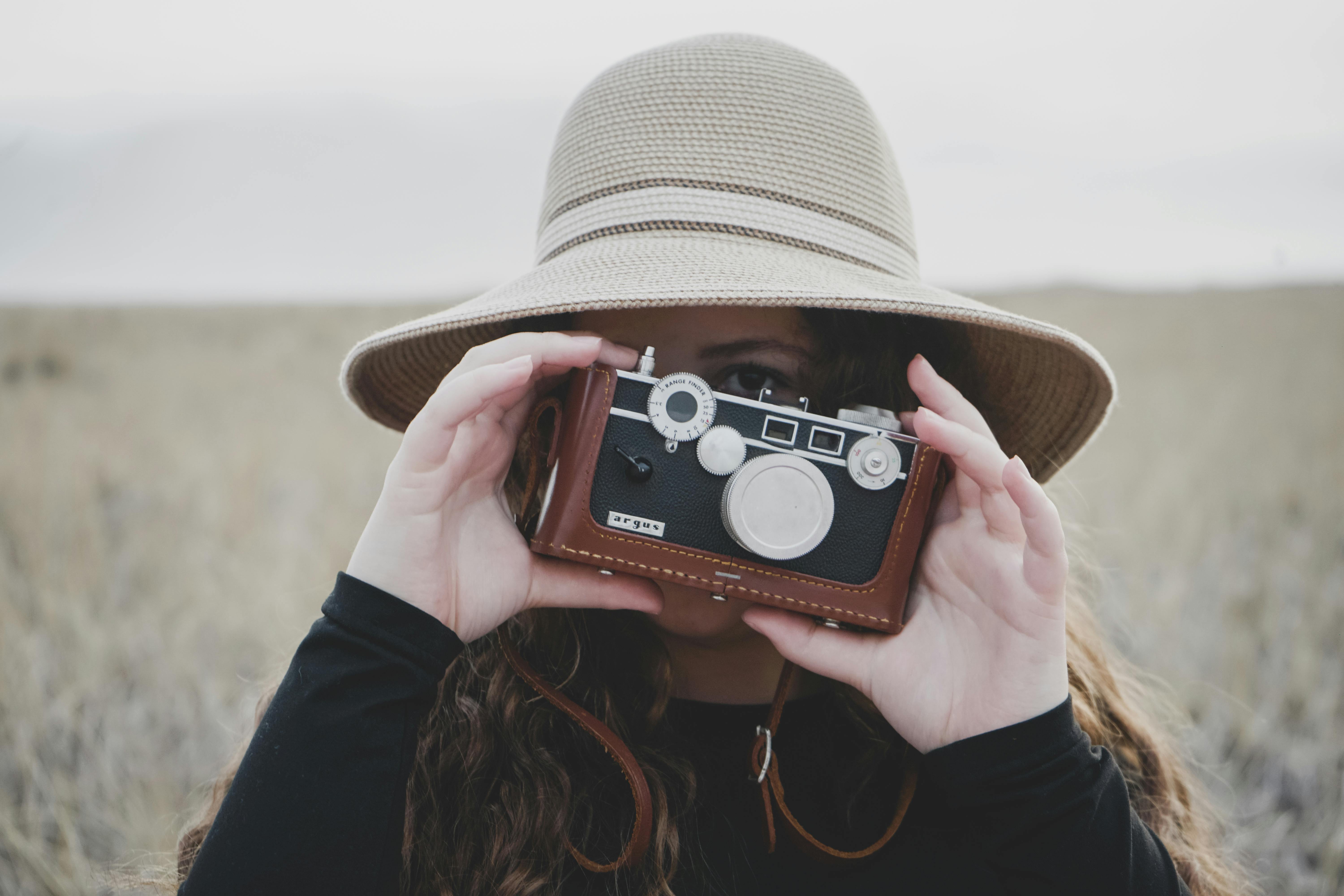 Woman wearing a wide-brimmed hat and black clothing, holding a vintage camera in front of her face in an outdoor setting.