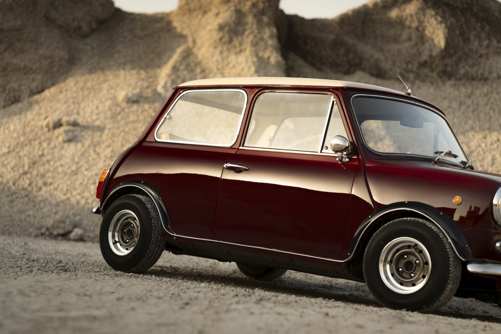 Classic maroon Mini Cooper on a gravel road with rocky hills behind.