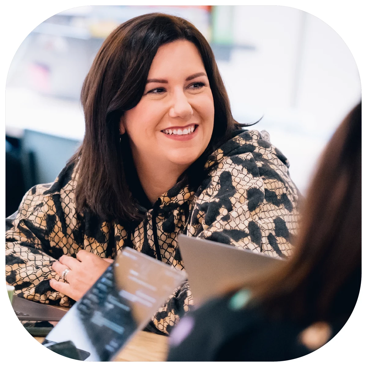 A woman with shoulder length dark hair is smiling at someone out of frame, she is wearing an animal print long sleeve shirt, there are laptops on the table in front of her