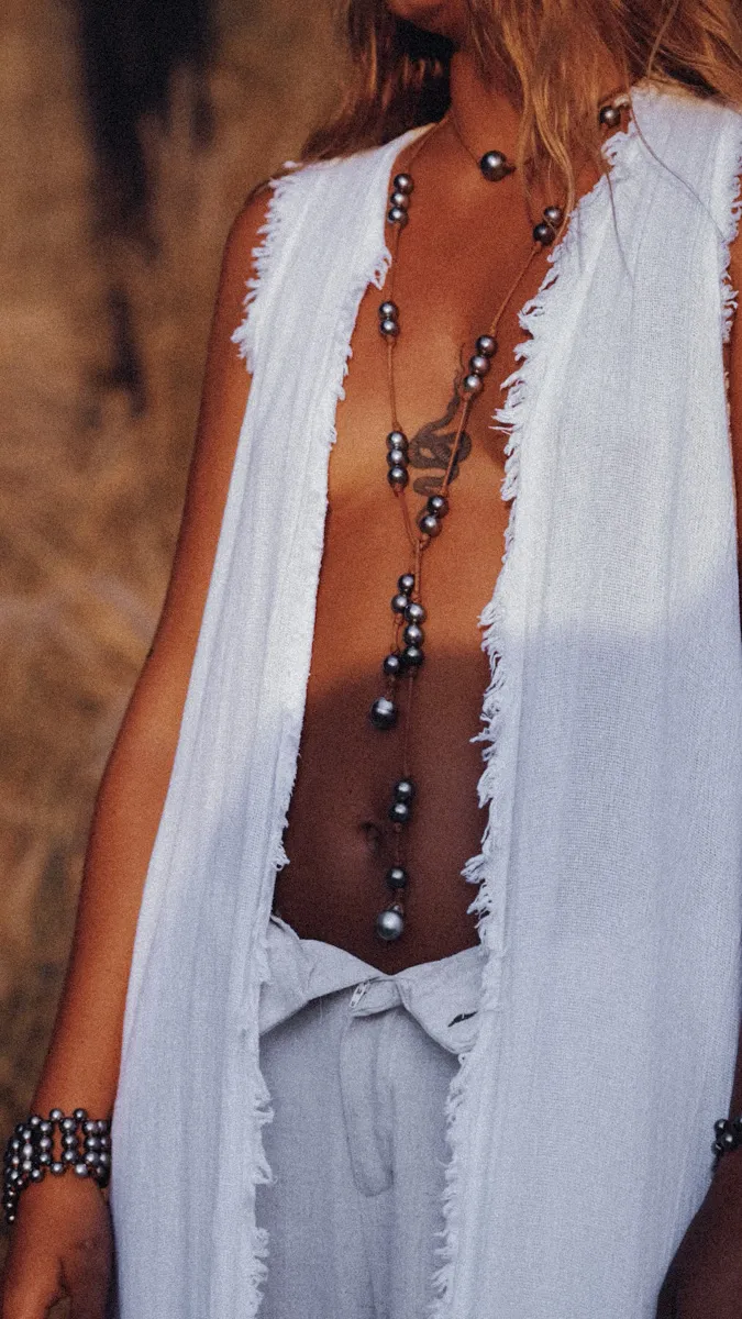 Woman in a frayed white linen vest layered with a long Tahitian pearl lariat necklace and a beaded pearl bracelet, deep grey pearls cascading along bare skin — effortless island luxury captured in golden light. Handmade luxury jewelry in Saint Barth — Manos de Oro Saint Barth.