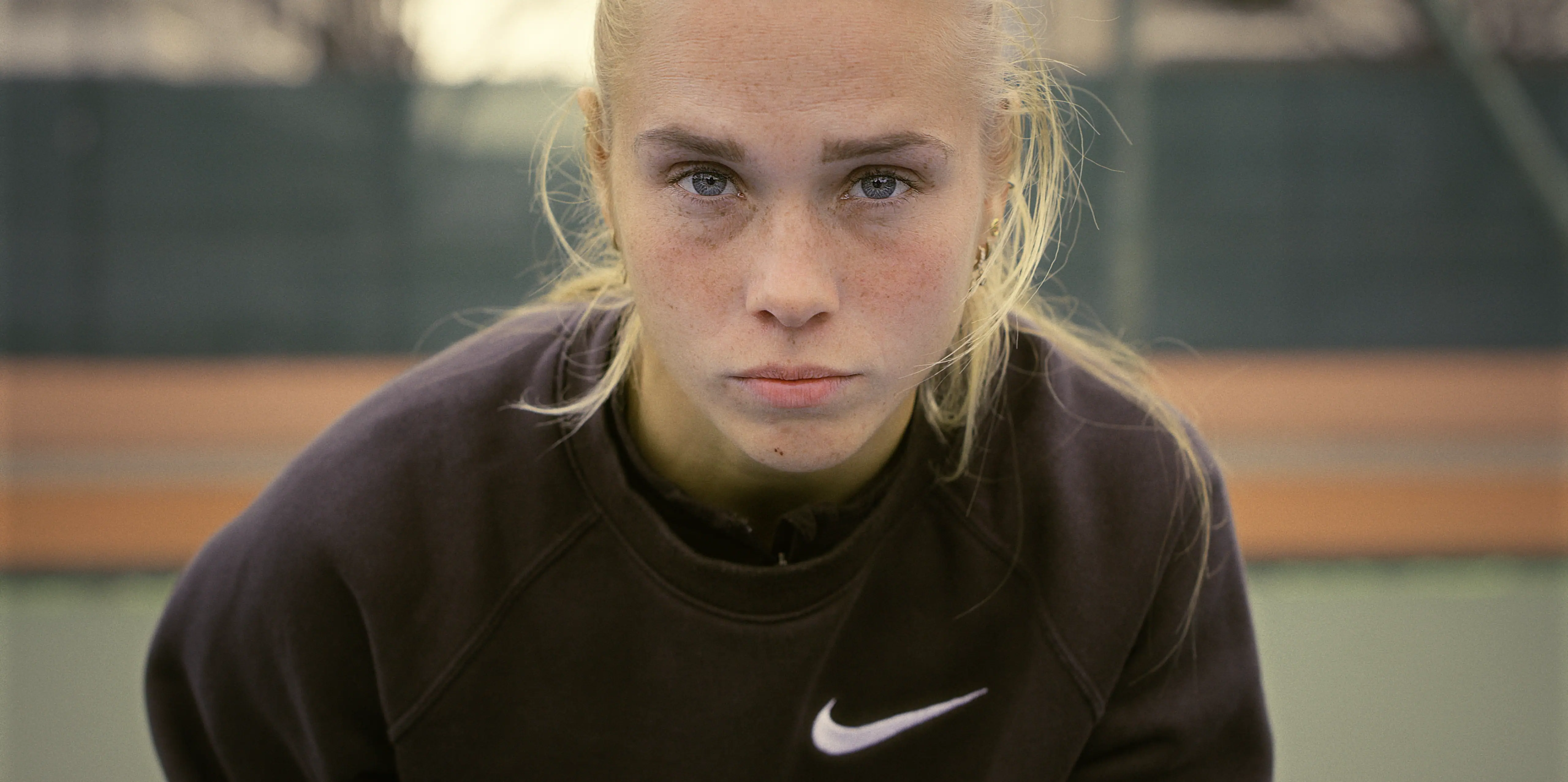 A female tennis player with blonde hair and a focused expression, wearing a black Nike sweatshirt, stands on a tennis court with a blurred background, signifying determination and athleticism.