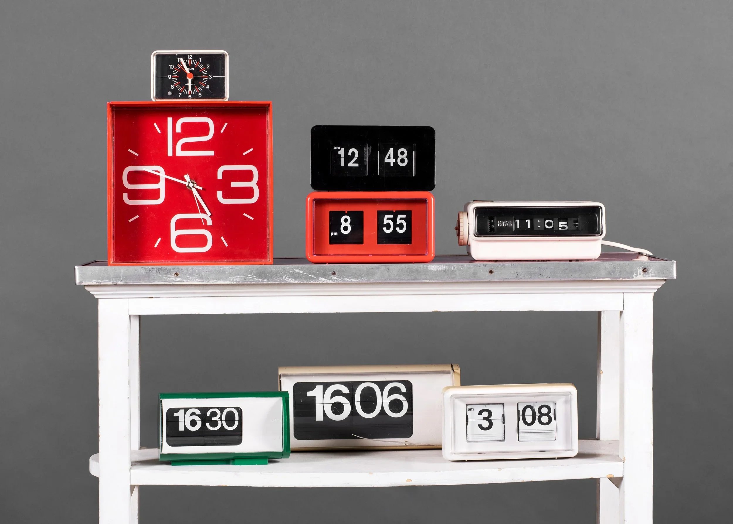 A collection of analogue and digital clocks arranged on a white stand against a grey wall.