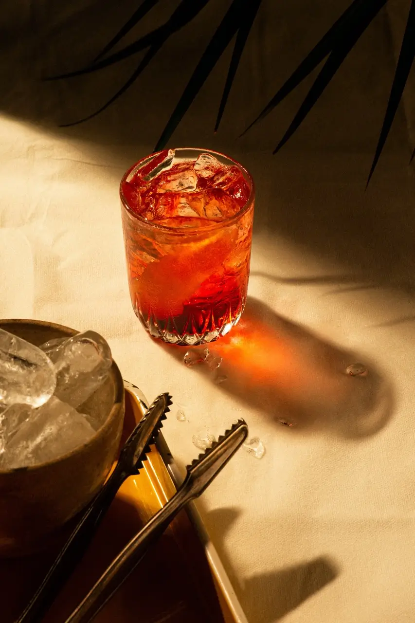 A crystal glass filled with a vibrant orange-red cocktail sits on a sunlit table, accompanied by a bowl of ice cubes and metal tongs, creating a refreshing and inviting scene.