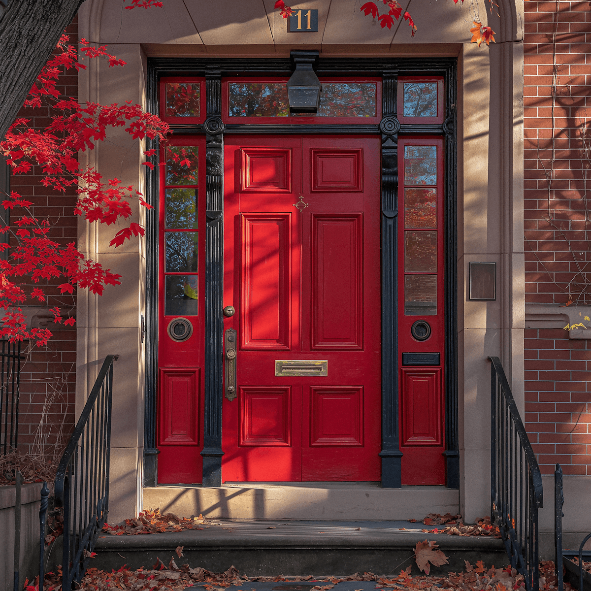 Classic brick townhouse entrance featuring a bold red panelled door, brass hardware, and framed by vibrant autumn foliage