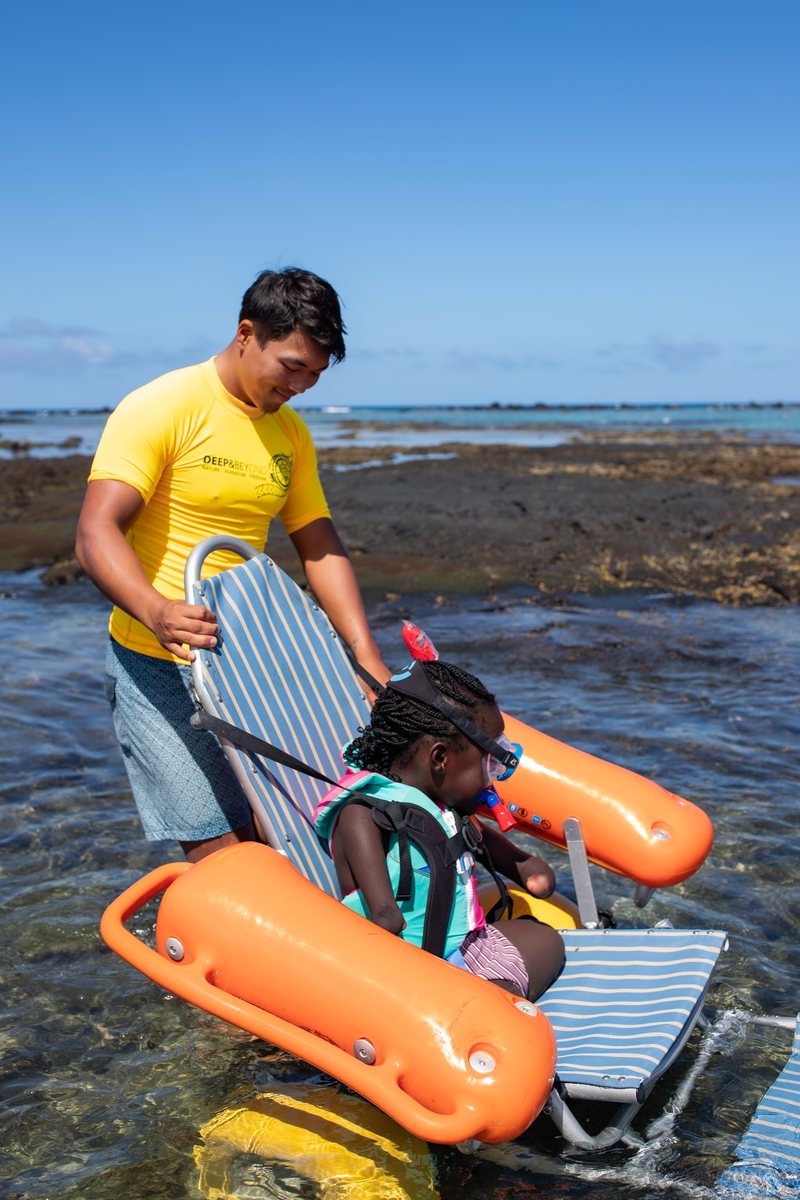 A volunteer guides a girl in an adaptive beach chair with orange floats into the water