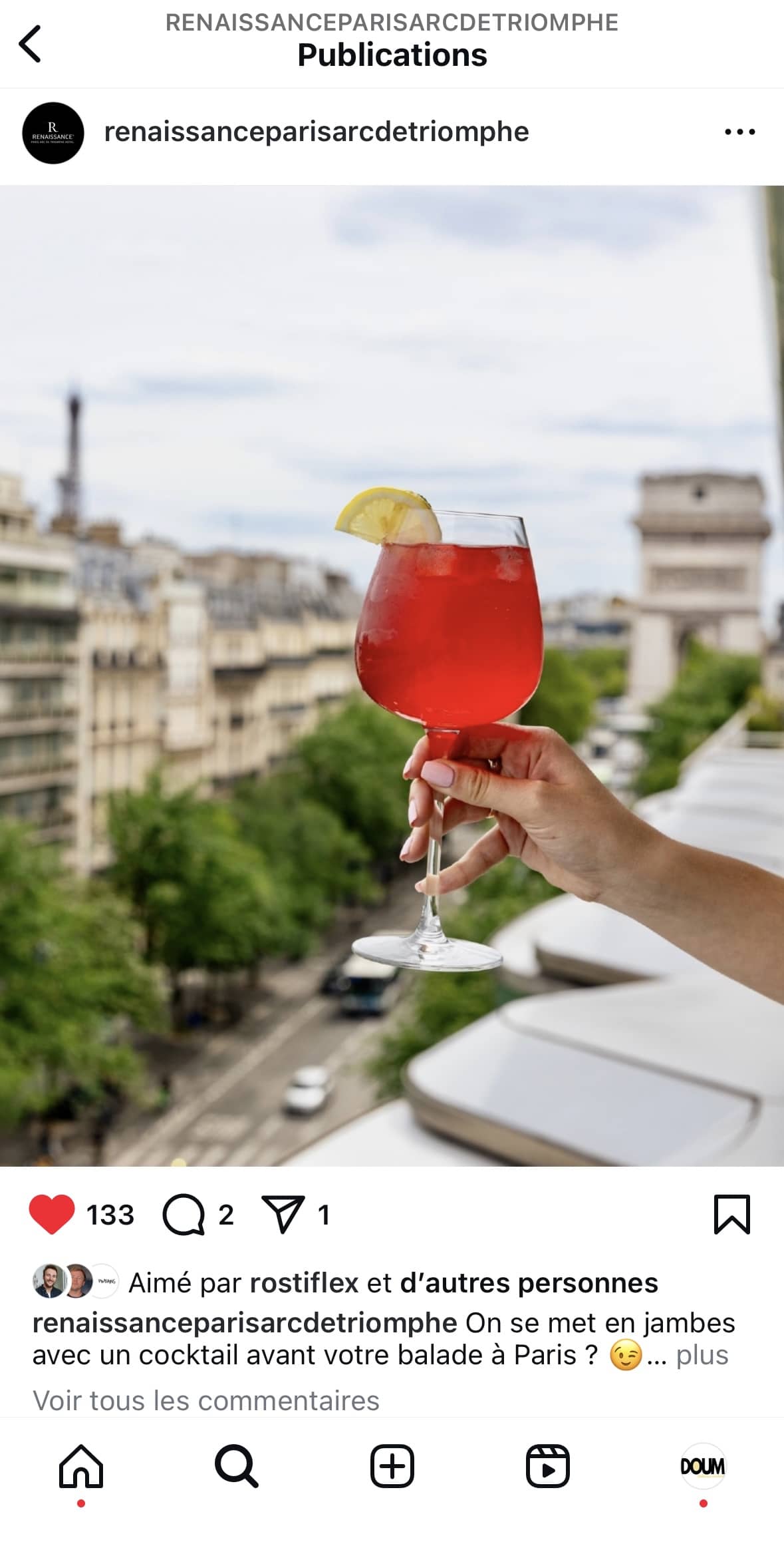 Un post instagram avec Une personne tient un grand cocktail rouge garni d’une tranche de citron sur un balcon avec vue sur les toits de Paris et la tour Eiffel en arrière-plan.