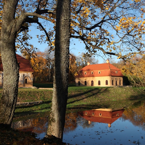 Two trees in the foreground, autumnal building with red roof reflected in a pond, surrounded by fall foliage and a clear sky.