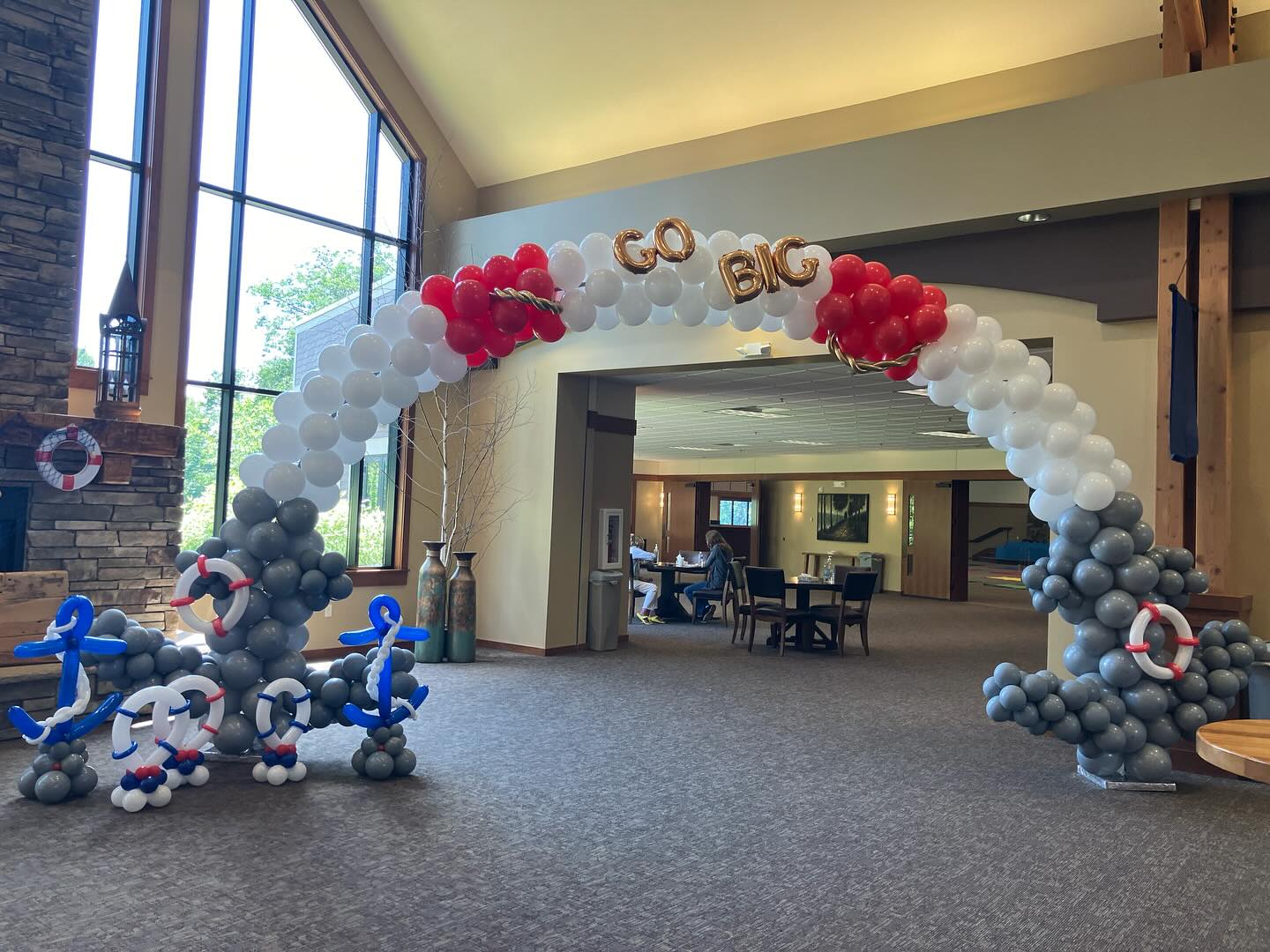 A large balloon arch in an office lobby features white, red, and gray balloons with the words "GO BIG" at the top, complemented by decorative anchors and lifebuoys.