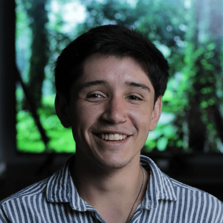 Woman in sunglasses poses against vibrant red background, wearing a beige blazer and black top. Sunlight casts dynamic shadows, creating a stylish, confident mood.