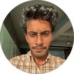 Young man with glasses in a gray hoodie, sitting in front of a computer screen with an orange background.