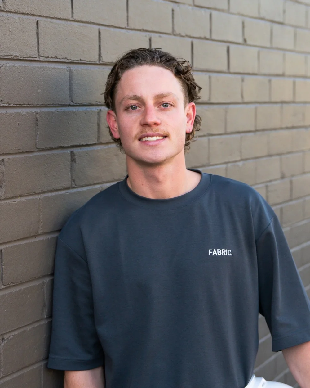 Man in gray t-shirt with "Fabric" logo leaning against a brick wall.