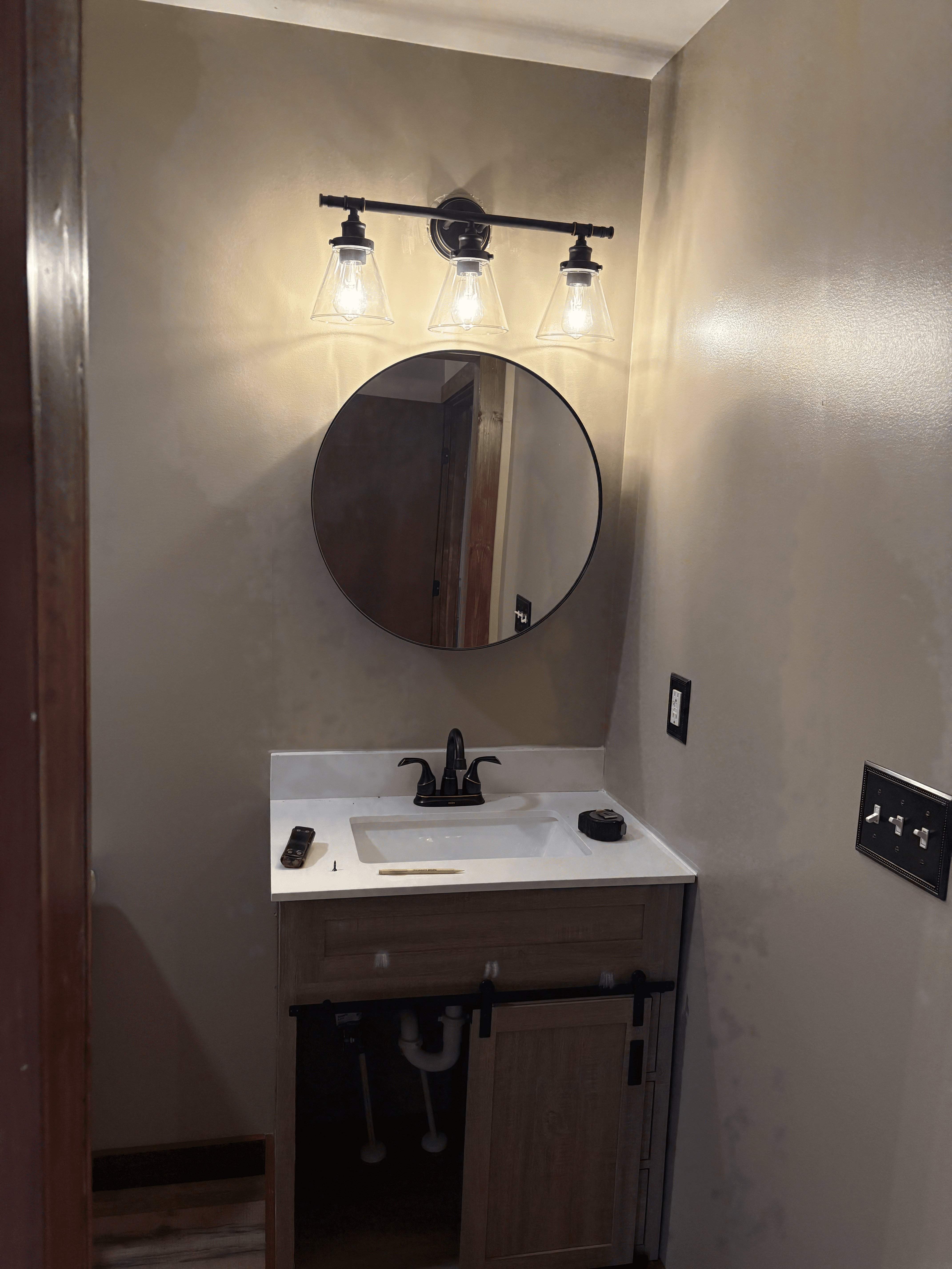 Professional bathroom vanity installation with quartz countertop, round mirror, modern lighting, black fixtures, and custom cabinetry.