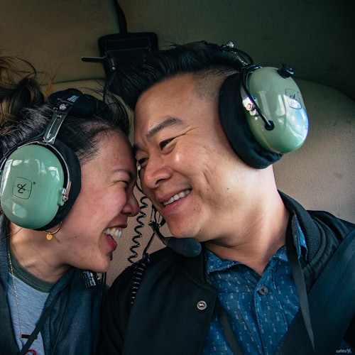 คนสองคนสวมชุดหูฟังสีเขียวและยิ้มให้กันอย่างใกล้ชิดภายในบริเวณที่ดูเหมือนเป็นห้องโดยสารของยานพาหนะหรือเครื่องบิน