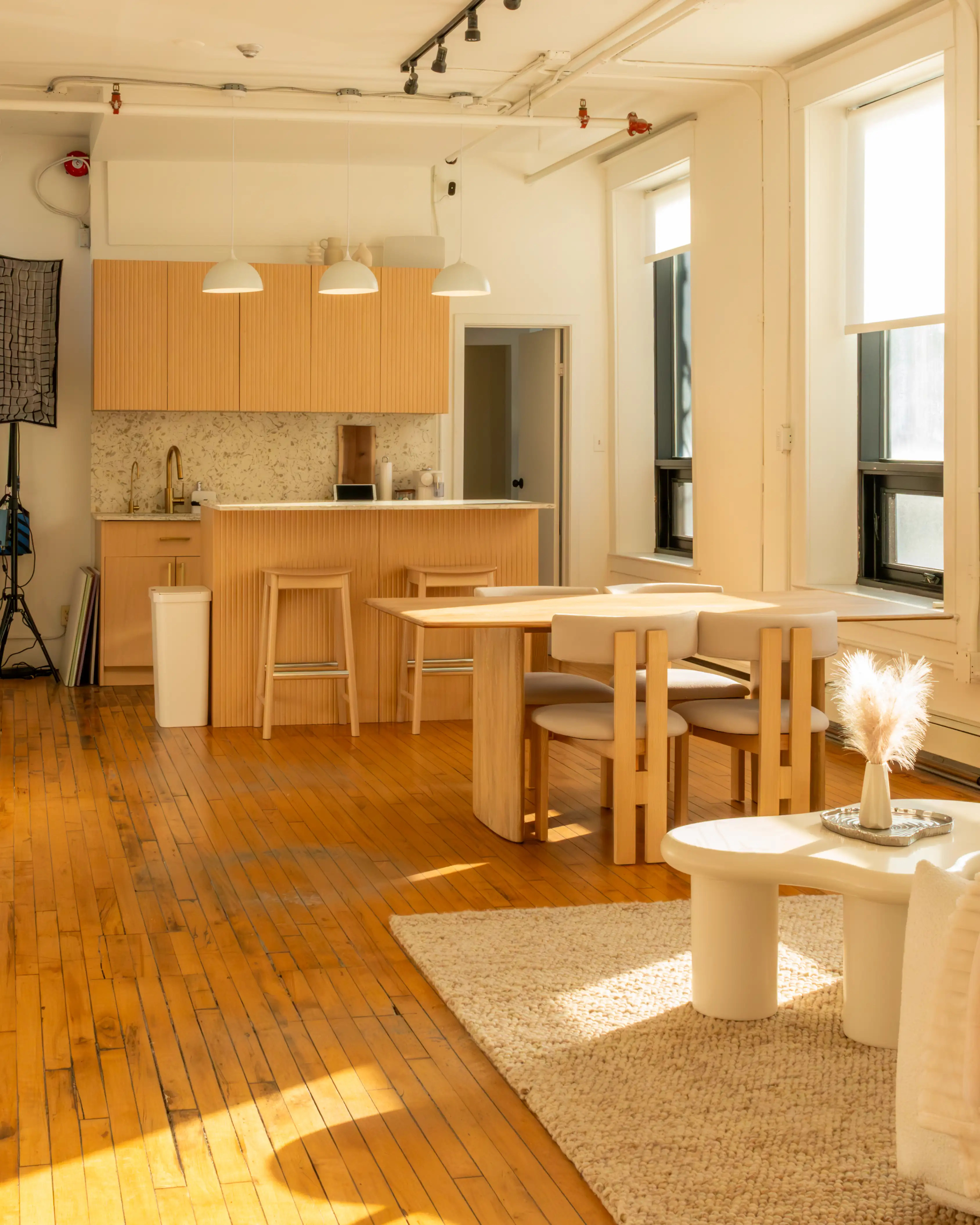 Node Studio’s semi-equipped kitchen with minimalist wooden cabinetry, a dining area with sleek furniture, warm hardwood floors, and natural light streaming through large windows, creating a versatile and inviting space