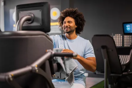 A smiling man with an afro exercises on a gym machine, surrounded by fitness equipment.