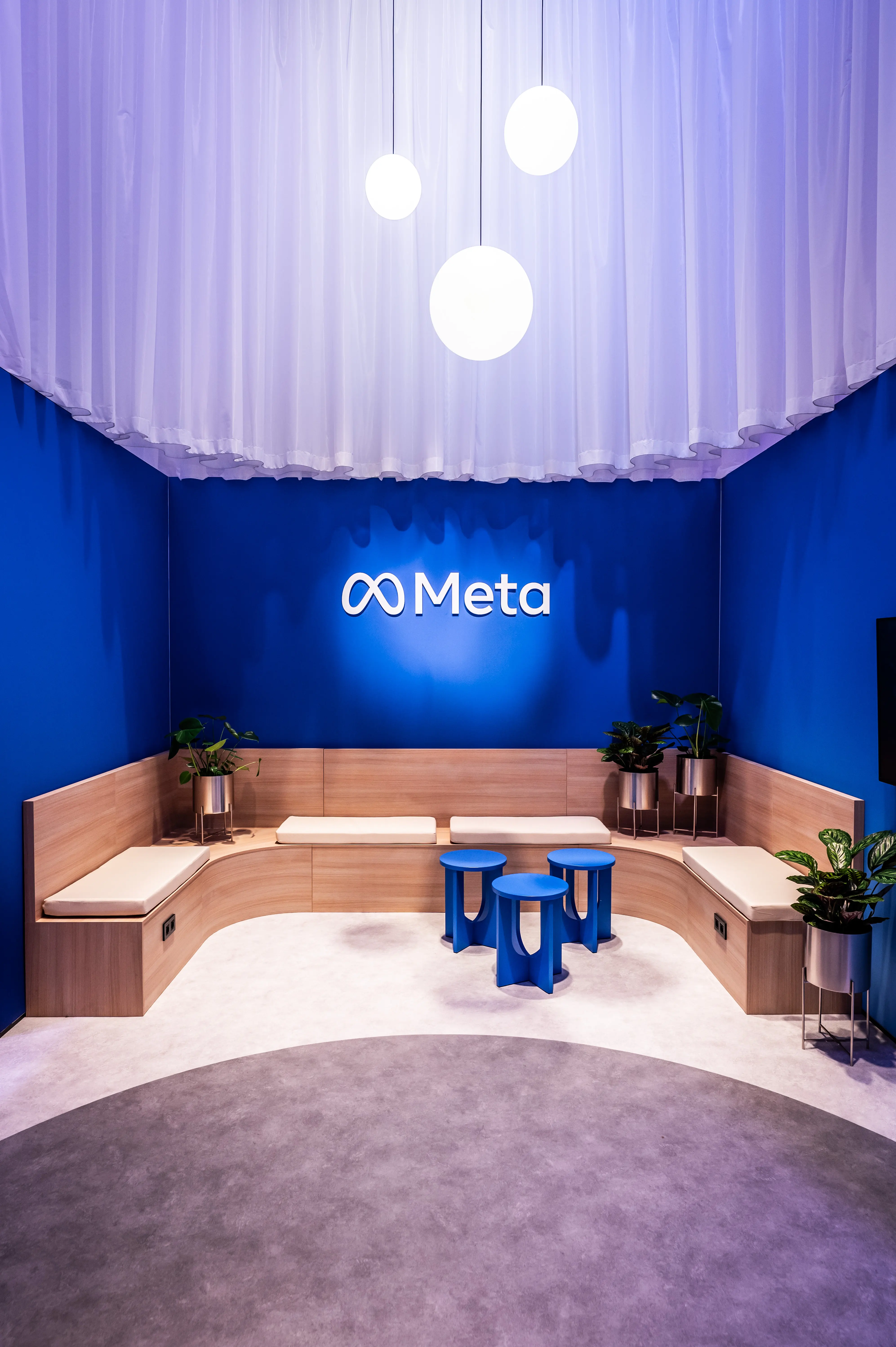 Blue Meta lounge with curved wooden bench, plants, blue stools and white curtain ceiling.