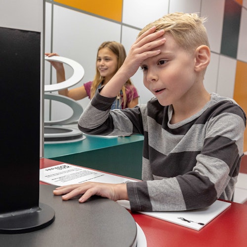 一個穿著條紋襯衫的男孩看起來困惑，撫摸著自己的額頭，背景中有一個微笑的女孩正在與一個展示互動。