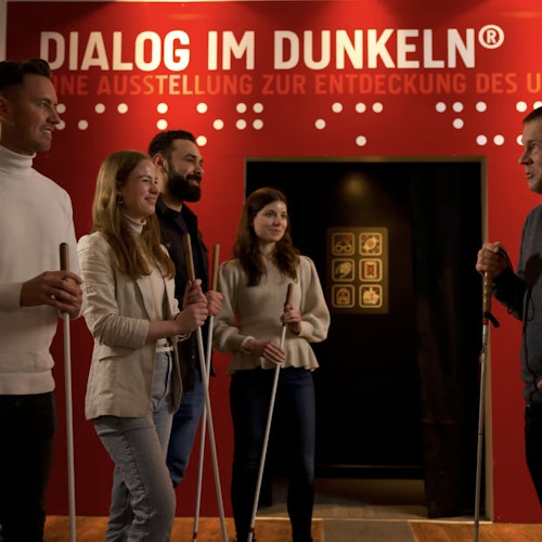 A group of five people stand in front of a red wall with the text "Dialog im Dunkeln." They are holding white canes.