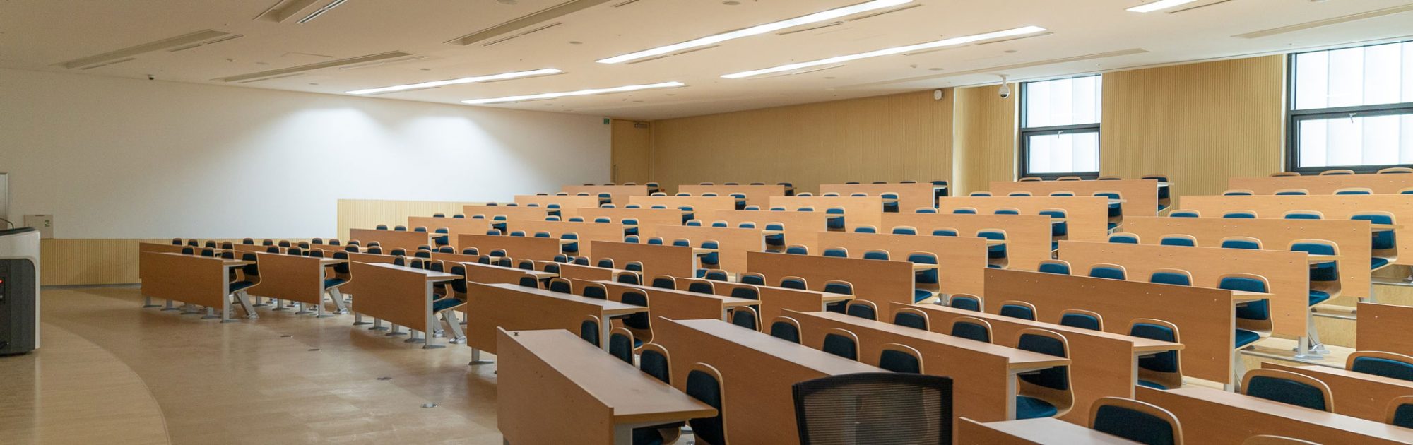 A spacious classroom with wooden desks arranged in rows, brightened by natural light from windows.