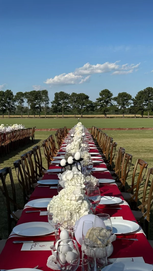 Mesa larga al aire libre con mantel rojo y arreglos blancos para celebraciones de gran escala que priorizan diseño escenográfico y coherencia visual.