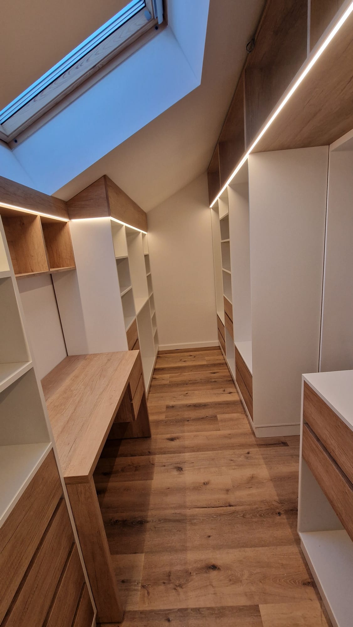 Dressing mansardă îngust, dulapuri albe și sertare din lemn, lumină Velux și LED discret.