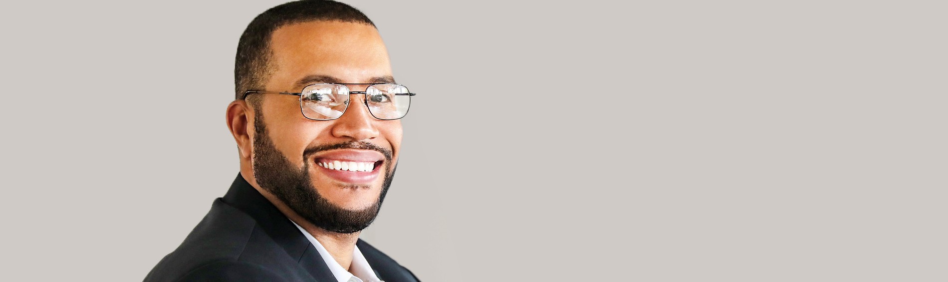 A smiling man wearing a suit, with glasses, against a neutral background. He appears friendly and approachable.