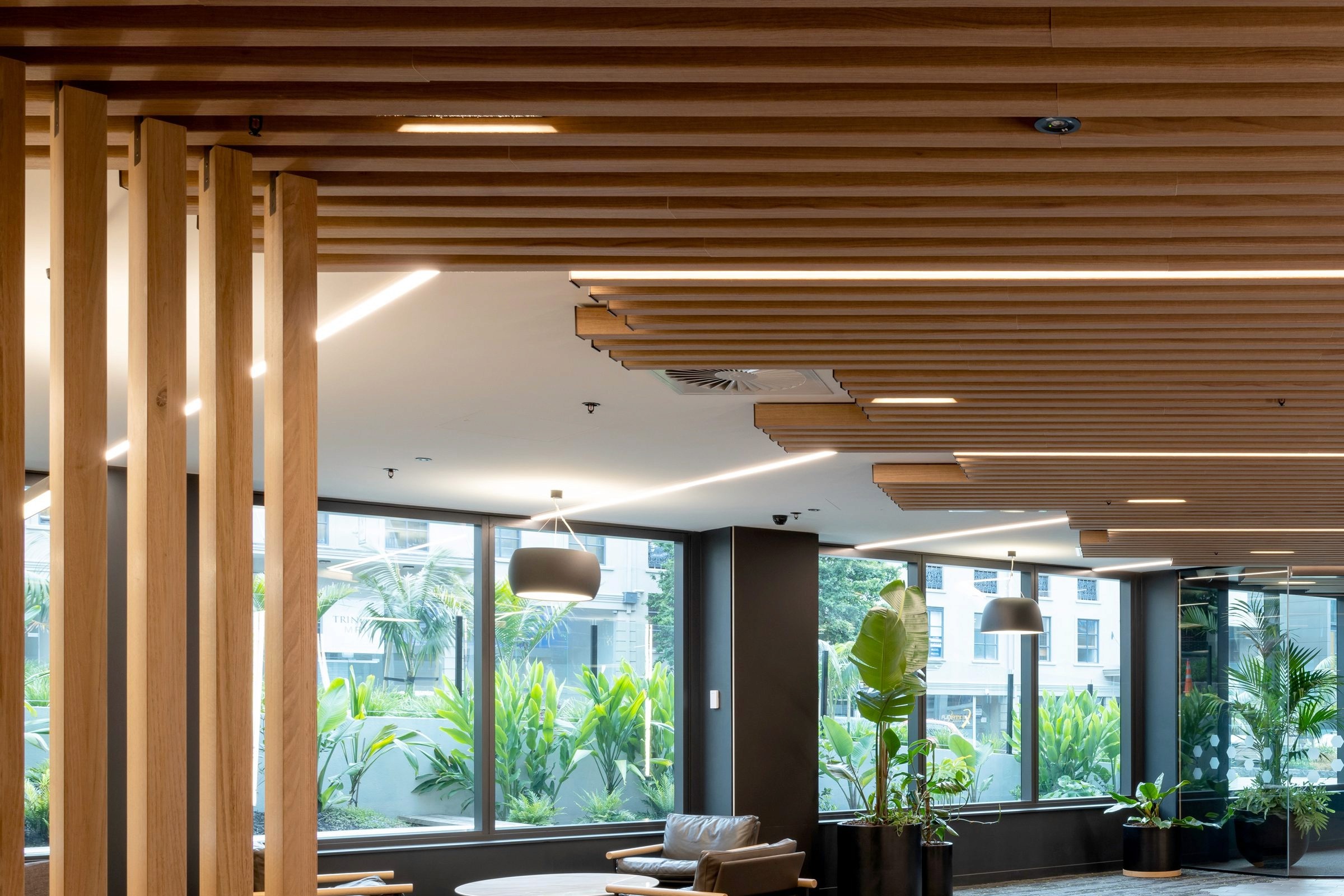 Modern lobby with a wood slat ceiling, linear lights, leather chairs, and large windows looking out at lush tropical plants.