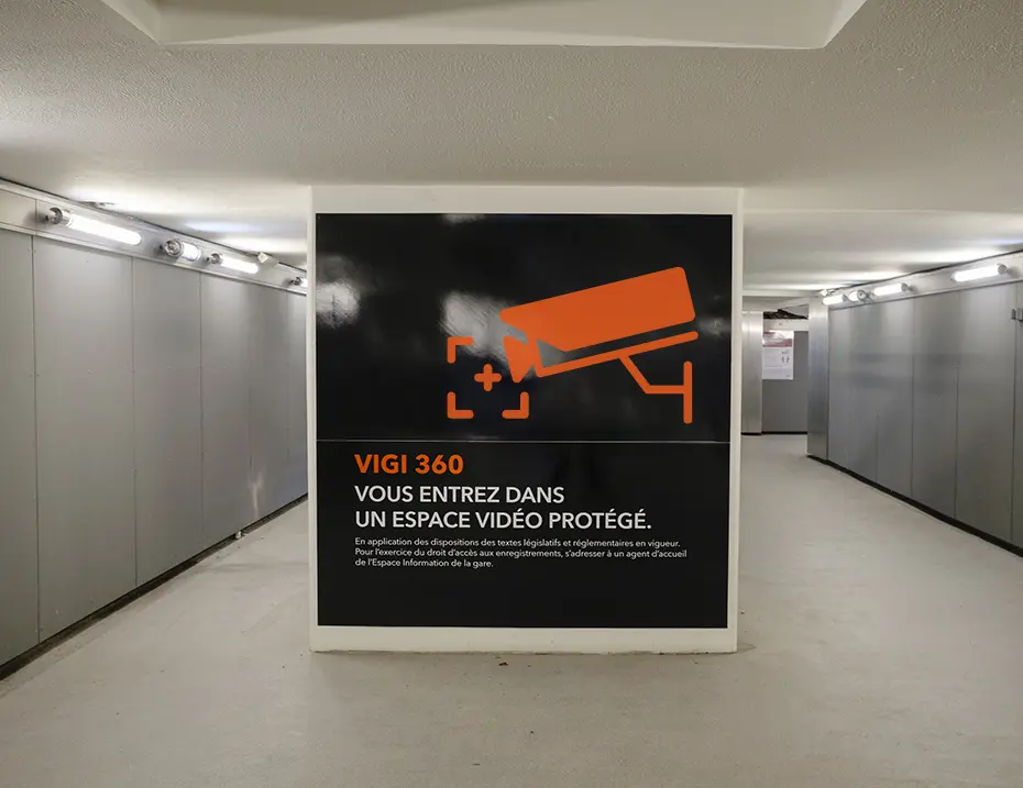 Large black wayfinding panel in an underground station corridor, displaying the orange camera pictogram and French text indicating entry into a video-monitored area.