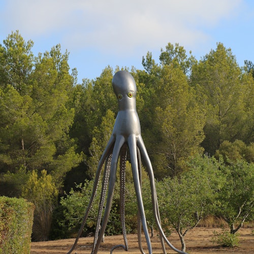 A large metallic octopus sculpture standing in front of a dense forest of green trees.
