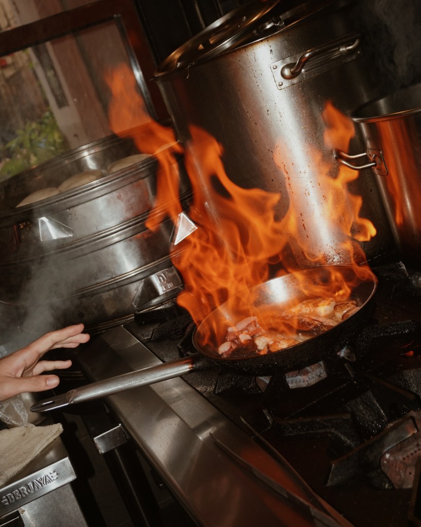 A chef's hand reaches towards a flaming pan on a gas stove, filled with sizzling ingredients, showcasing an intense cooking moment in a restaurant kitchen.