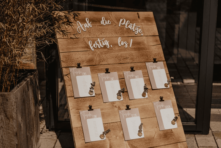 Kreativer Sitzplan auf einem Holzbrett mit der Aufschrift 'Auf die Plätze, fertig, los!' und angeklemmten Kärtchen mit Eukalyptus und Wachssiegeln.