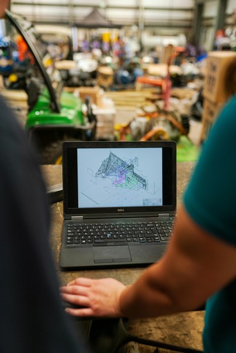 Laptop su banco da lavoro mostra un progetto 3D CAD, con persone e macchinari sfocati sullo sfondo dell’officina.