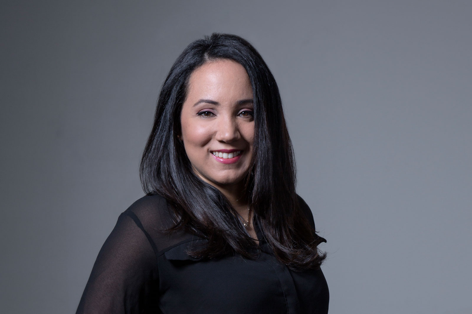 A woman with long, dark hair is captured in a head-and-shoulders portrait against a solid gray background. She is wearing a black top with sheer sleeves and a subtle sheen. Her face is lit up with a warm smile, and she gazes directly at the camera. The lighting is even and soft, highlighting her features without harsh shadows.