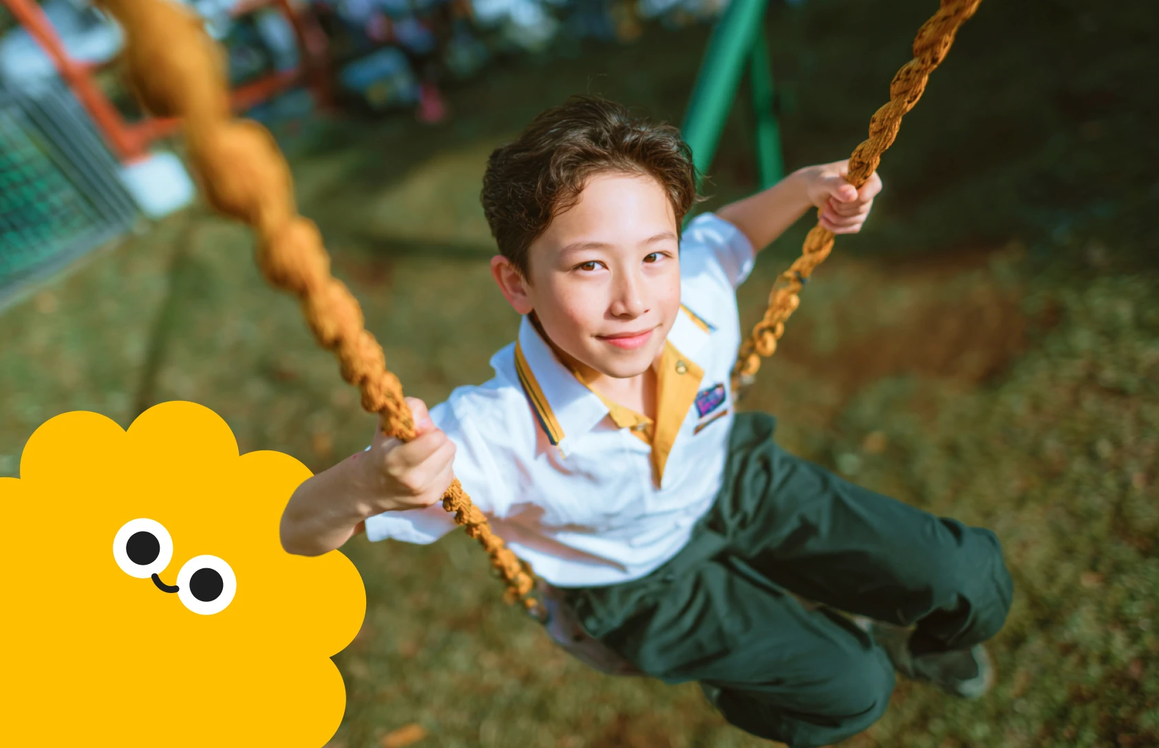 A young child smiles while swinging on a rope swing in a sunny playground. The child wears a white shirt and green pants. A yellow cartoon cloud smiles in the foreground, adding a playful tone.