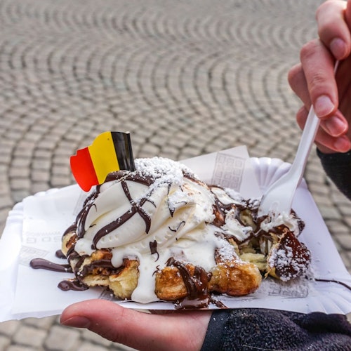 Ruka koja drži papirnati tanjur s vafelom prelivenim sladoledom, čokoladnim umakom, šećerom u prahu i malom belgijskom zastavicom.