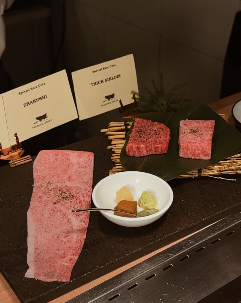 A selection of premium beef cuts, including Shakushi and Thick Sirloin, presented on a black stone surface alongside dipping sauces and garnishes.