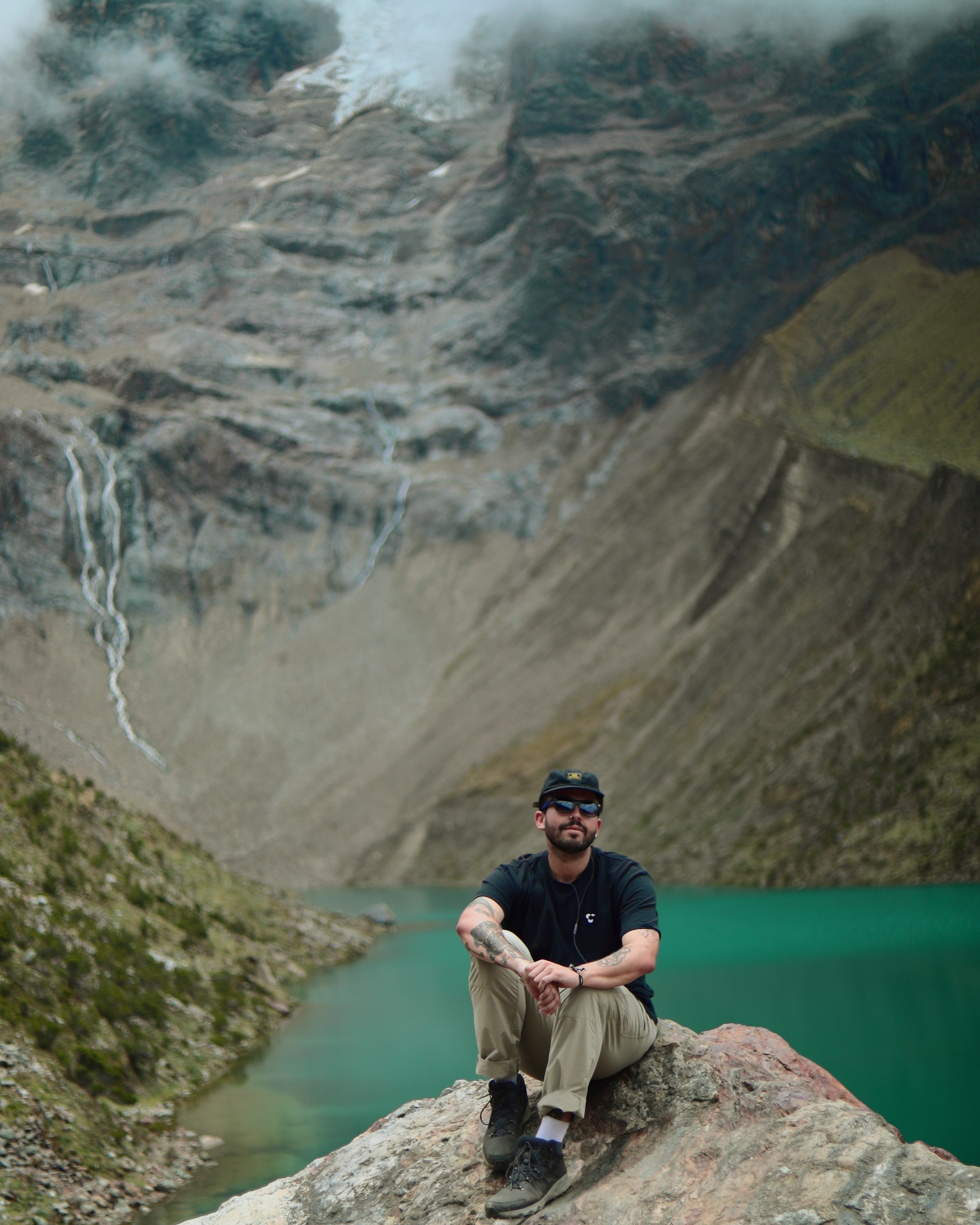 A man sits on a rock next to a lake with his knees drawn up to his chest, looking at the camera