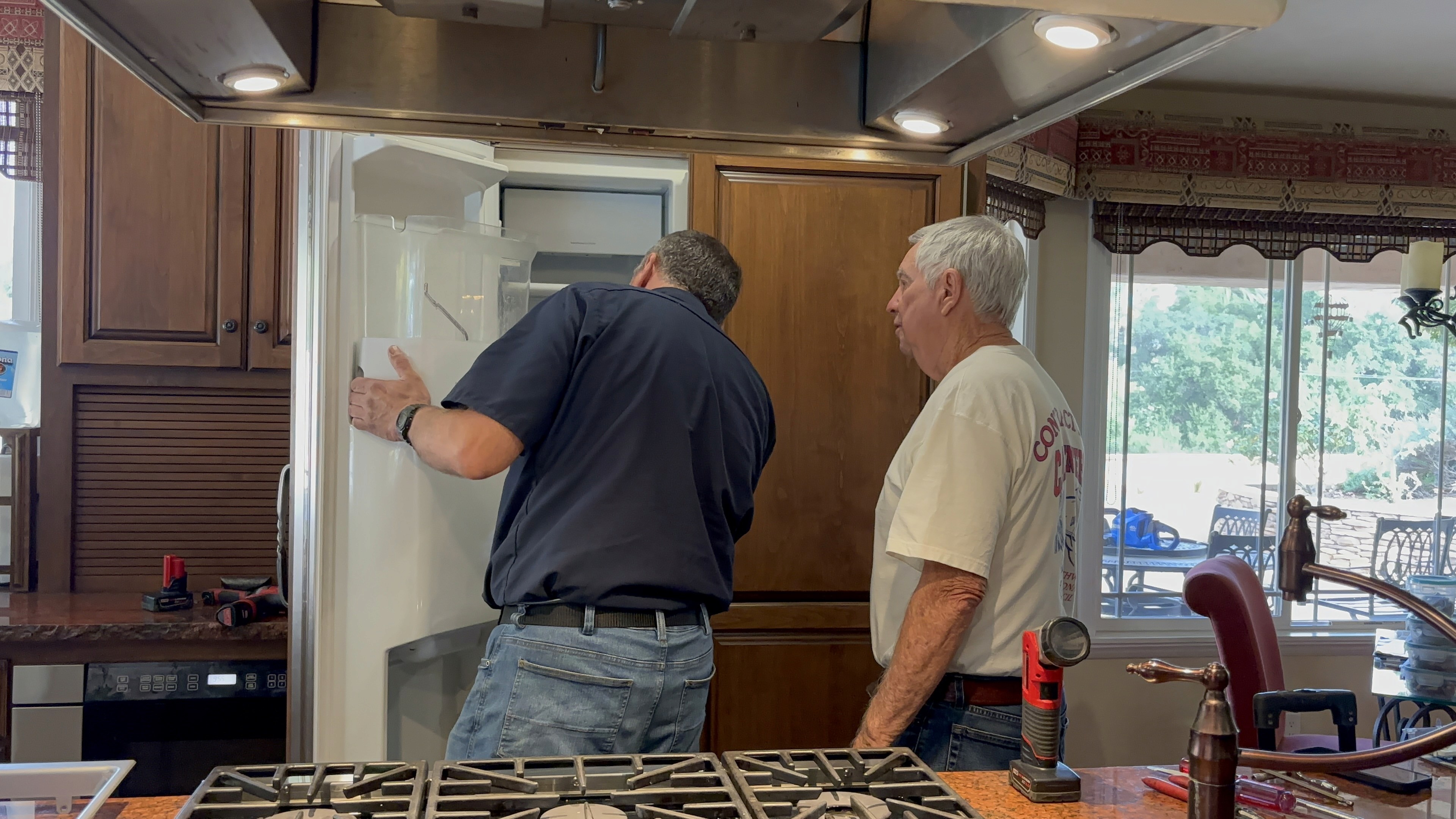 An older man, around 50 years old, dressed in a technician's uniform with 'AngelSky' branding, working on a refrigerator in a typical American kitchen