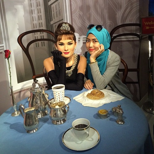 A woman in a blue headscarf poses with a wax statue of Audrey Hepburn at a table set with breakfast items and silverware.