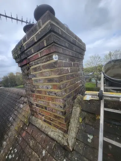 Old brick chimney stack with visible moss and weathering on a pitched tiled roof in the UK.