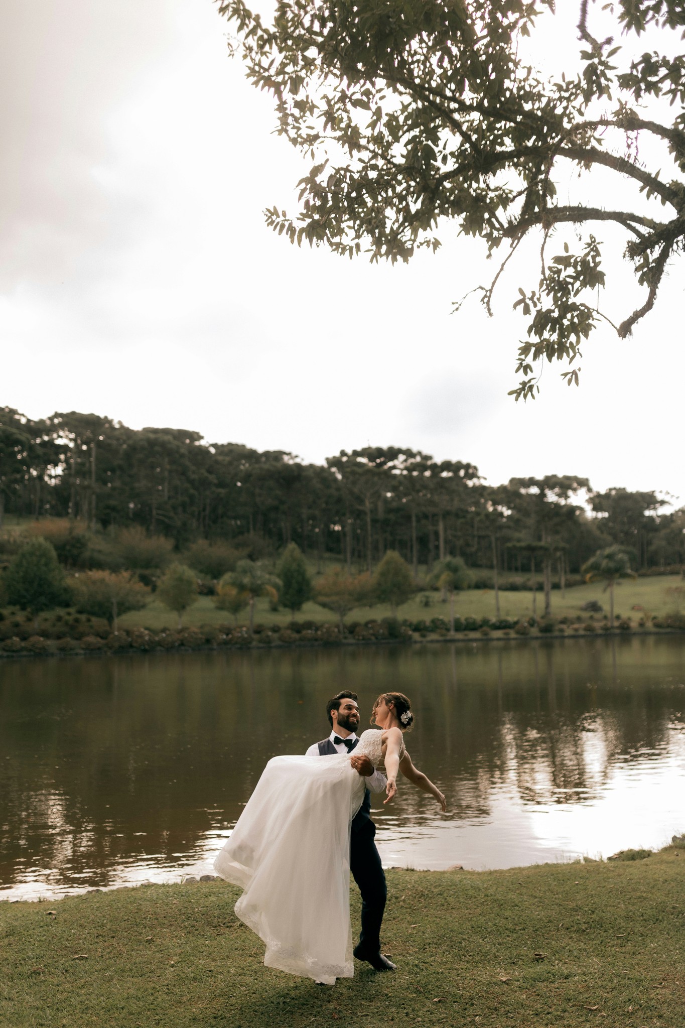 Groom lifting bride in his arms by a lake with rolling green hills and trees in the background