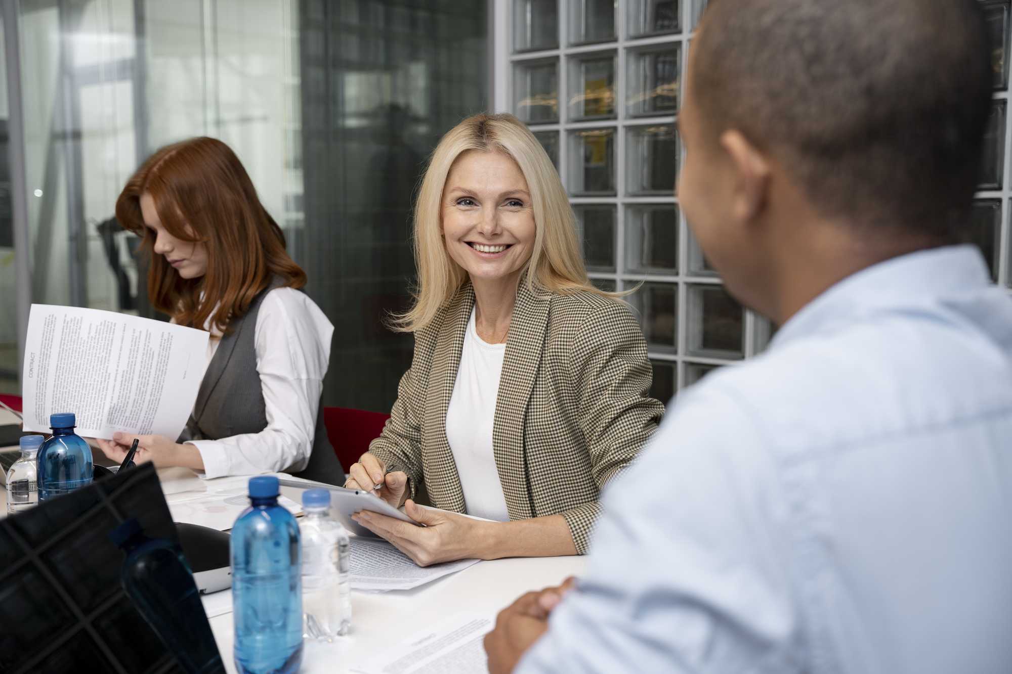 A blonde woman smiling and holding a tablet during a meeting, with colleagues engaged in reviewing documents around her