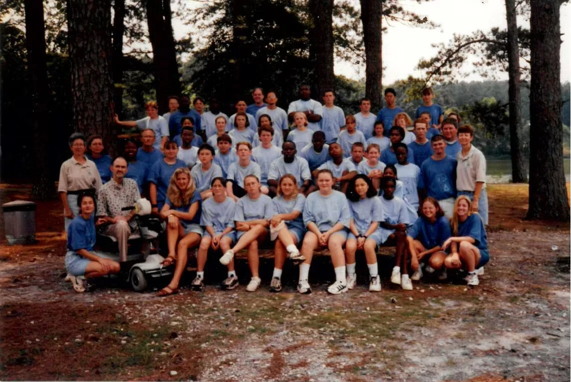 Large group photo of campers and staff in light blue Camp Juliena shirts, posing outdoors in a wooded area. Some are seated on a bench.