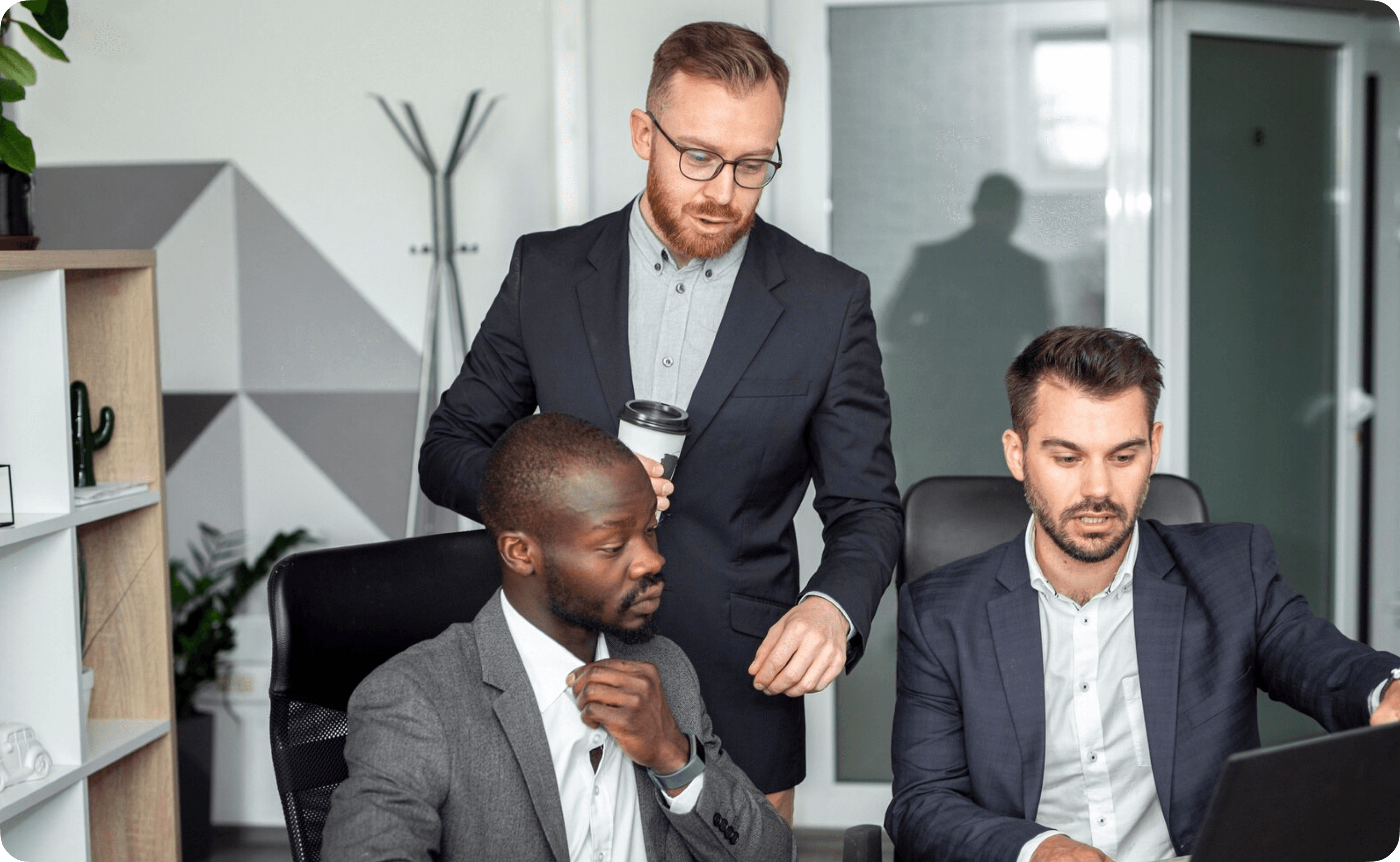 Three people in business attire discussing around a laptop in a modern office.
