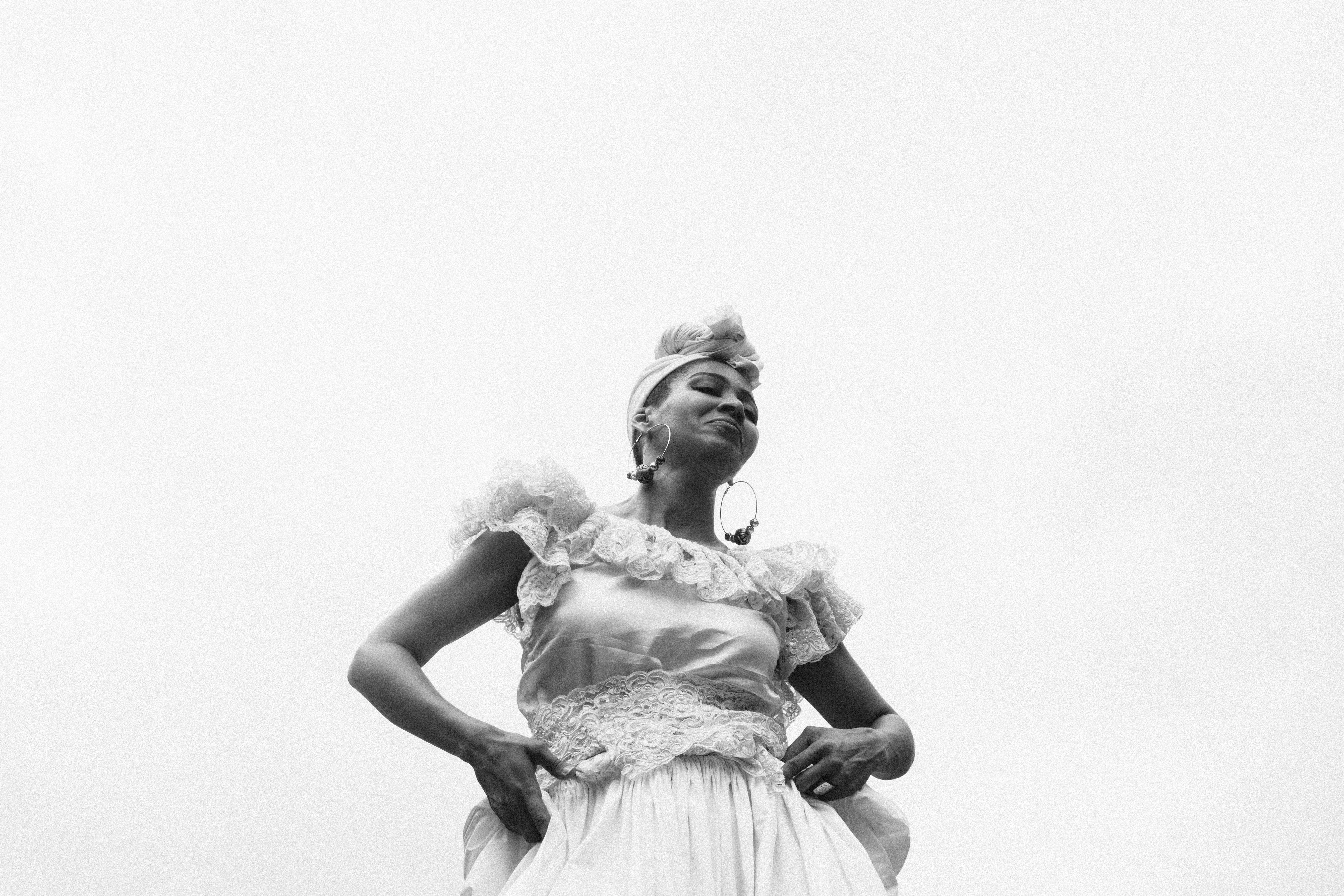 A person wearing a traditional, ornate dress with lace details stands confidently against a cloudy sky, exuding a sense of pride and cultural heritage.