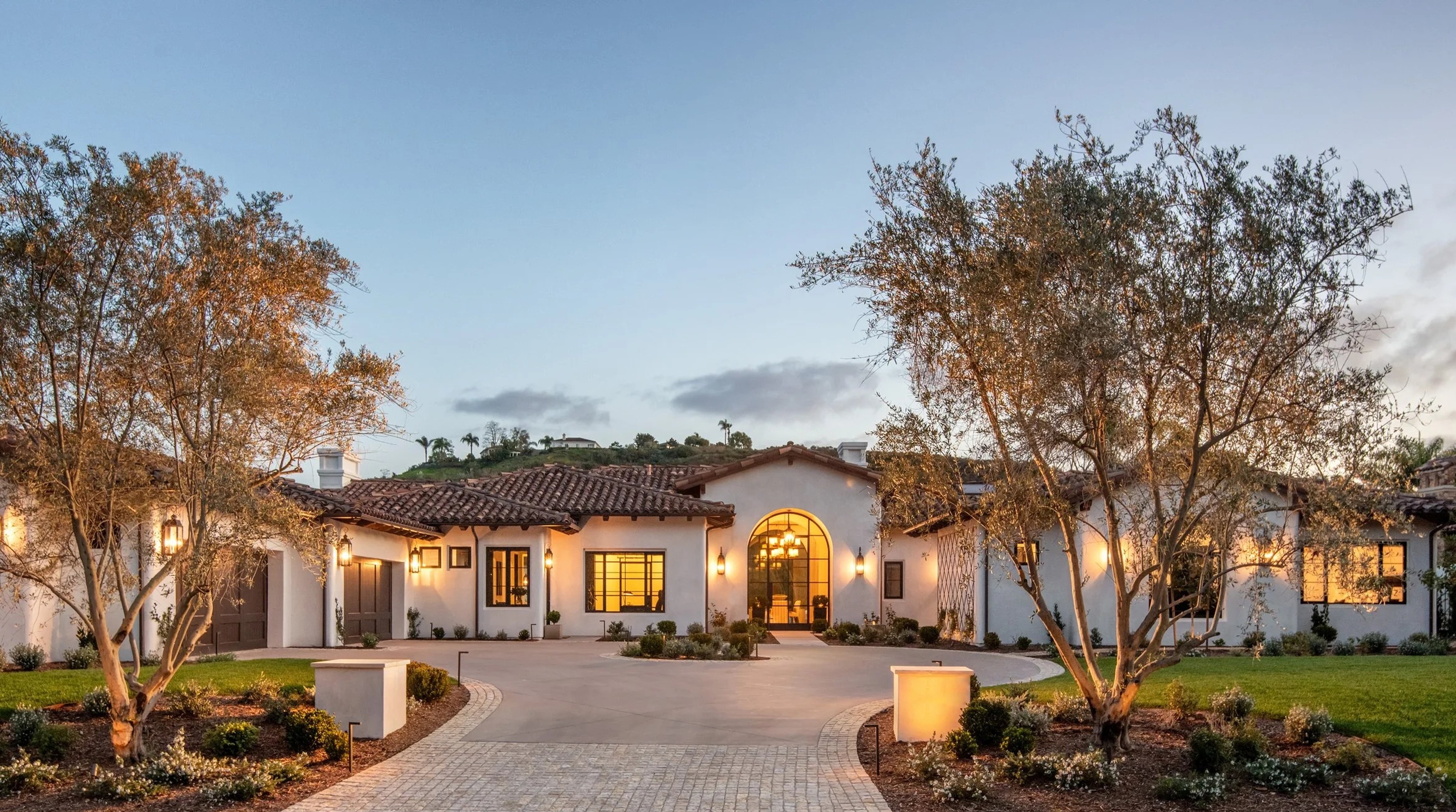 Front exterior of a luxury Spanish-style villa at dusk with white stucco walls, terracotta tile roofs, and arched entryways. Illuminated windows and outdoor lanterns cast a warm glow on a paved circular driveway and olive trees.
