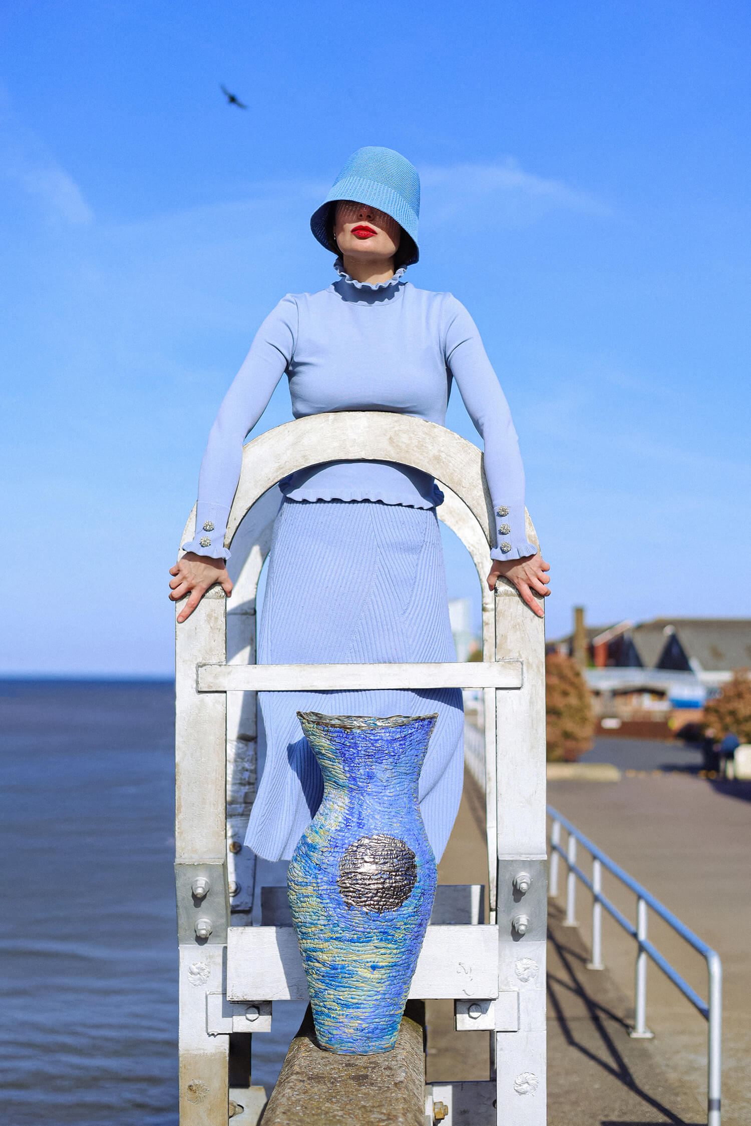 Fashion portrait of woman in blue outfit and bucket hat standing on coastal pier.