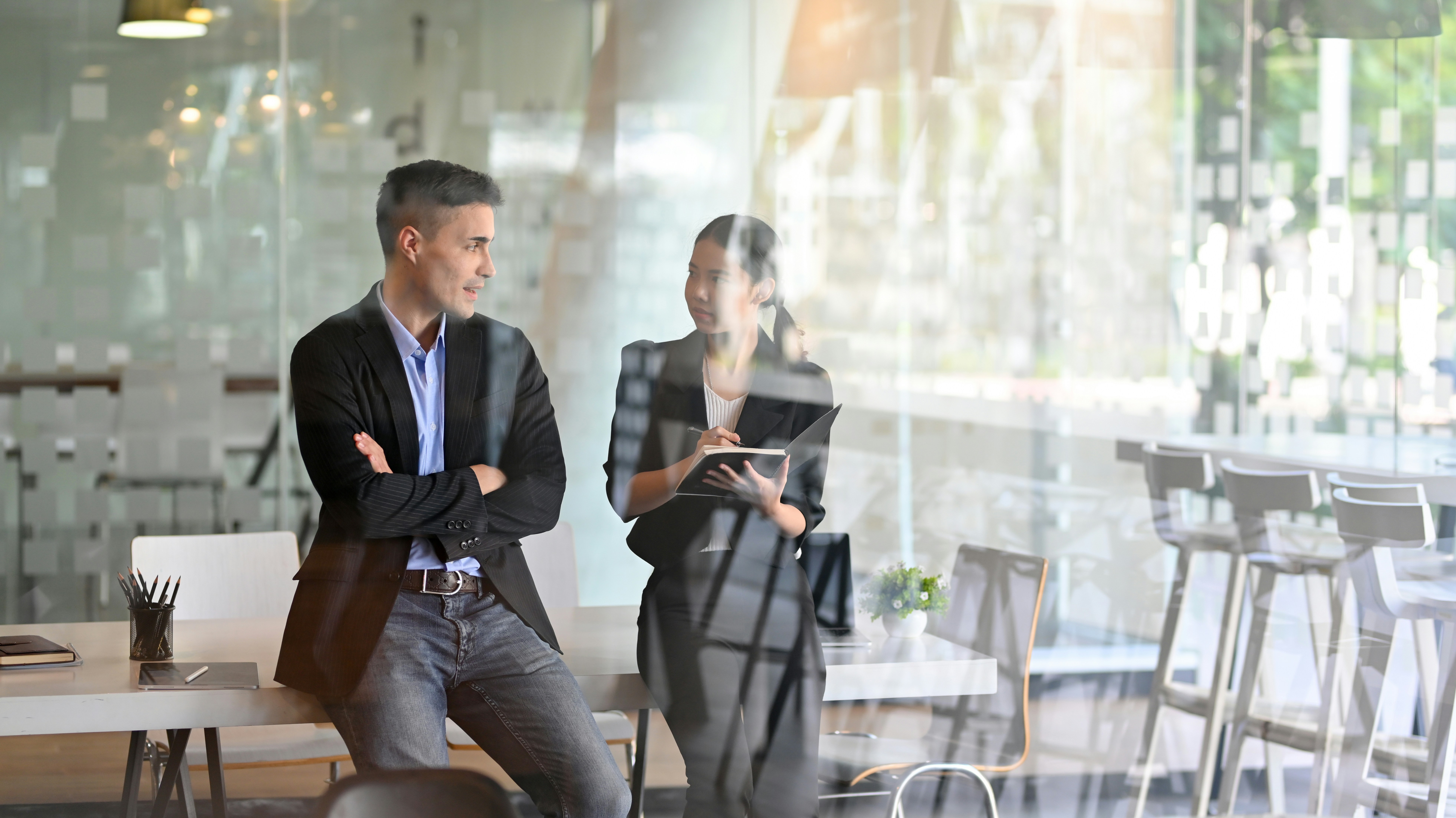 Two professionals seated in a modern office space, engaged in discussion, with large windows in the background.