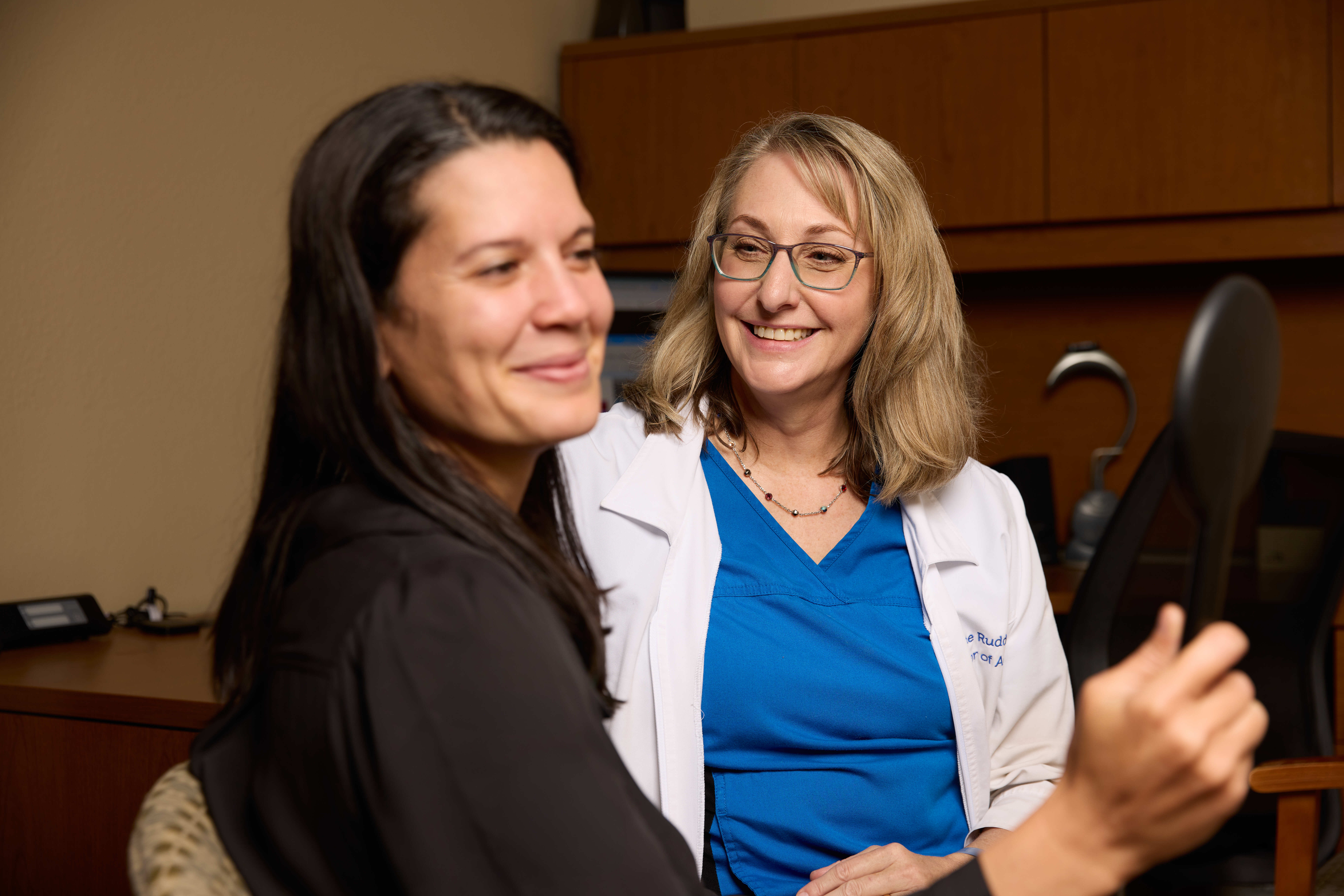 Two smiling women, one in a blue shirt and the other in a black dress, share a joyful moment together.